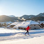 Skieur de fond en rouge sur une piste de Corrençon en Vercors, avec des chalets enneigés et montagnes boisées sous un ciel clair.