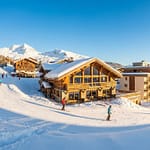Trouver le logement à La Rosière idéal pour votre séjour Vue panoramique de la station de ski La Rosière avec skieurs, chalets en bois, résidences modernes et montagnes enneigées sous ciel bleu.