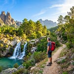 Aiguilles de Bavella : randonnées et cascades en Corse Un randonneur en Corse devant une cascade et une piscine naturelle aux pieds des Aiguilles de Bavella et d'une forêt luxuriante.