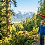 Famille en randonnée sur un sentier boisé. Le père pointe des montagnes enneigées au loin, la mère tient une carte. Enfants émerveillés.