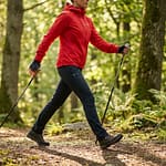 Femme pratiquant la marche nordique sur un sentier forestier ensoleillé, vêtue d'une veste rouge et utilisant des bâtons.