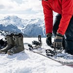 Gros plan sur une personne ajustant son snowboard et ses fixations, bottes et casque à proximité, avec un fond de montagnes enneigées.