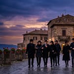 Neuf personnes élégantes en manteaux et chapeaux noirs se promènent sur une rue pavée d'une ville historique italienne au crépuscule.