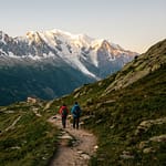 Deux randonneurs marchent sur un sentier rocailleux vers un refuge de montagne, avec les sommets enneigés du Mont Blanc illuminés par le soleil levant en arrière-plan.