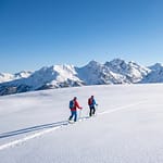 Deux skieurs de randonnée progressent sur un plateau enneigé. Des montagnes imposantes dominent l'horizon sous un ciel bleu limpide.
