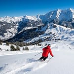 Skieur en veste rouge sur une piste immaculée, avec les sommets enneigés et forêts des Alpes sous un ciel bleu clair.