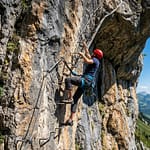 Homme en casque rouge escaladant une via ferrata rocheuse. Paysage alpin avec vallées vertes et ciel bleu à l'arrière-plan.