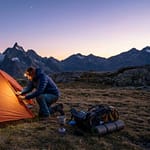 Réussir son premier bivouac : tout ce qu'il faut savoir Randonneur préparant sa tente orange au coucher du soleil face à des sommets enneigés.