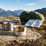 Un réchaud à gaz et un réchaud à alcool posés sur une roche en montagne avec un sac à dos et une carte.