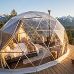 Séjour glamping dans les Alpes : le luxe en pleine nature Un dôme géodésique transparent avec un lit et un jacuzzi en bois sur une terrasse surplombant des montagnes enneigées.