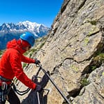 Personne équipée d'un casque bleu et d'une veste rouge pratiquant la via ferrata face au massif du Mont-Blanc.