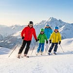 Les stations de Savoie idéales pour skier en famille Une famille souriante skie ensemble sur une piste ensoleillée avec les montagnes enneigées en arrière-plan.