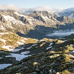 Un randonneur portant une veste bleue et un sac à dos marche sur un sentier face à un panorama de sommets enneigés.
