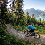 Parcourir Annecy en VTT : 5 circuits incontournables Vététiste en maillot bleu roulant sur un sentier forestier avec vue panoramique sur le lac d'Annecy et les montagnes.