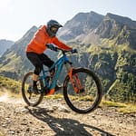 VTT électrique en montagne : les meilleurs modèles 2026 Un vététiste en équipement orange sur un VTT électrique bleu descendant un sentier rocheux en montagne.