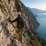 Randonneur de dos progressant sur une via ferrata surplombant un lac alpin turquoise